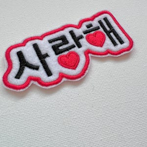 May include: A white embroidered patch with red stitching and two red hearts. The patch says "사랑해" in black text.