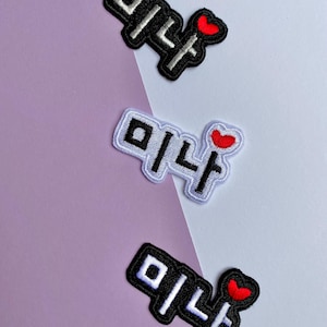 Könnte beinhalten: Drei gestickte Aufnäher mit dem koreanischen Wort "미나" in weißen Buchstaben mit schwarzem Rand, jeweils mit einem kleinen roten Herz. Die Aufnäher sind auf einem lila und weißen Hintergrund angeordnet.