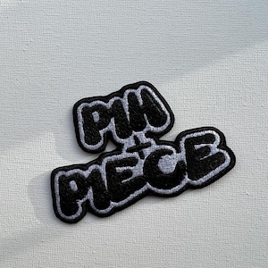 May include: Black and white embroidered patch with the text "PIH + PIECE" in a bubble letter style. The patch has a black border and is set against a white background. The patch is likely for clothing or accessories.