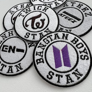 Può includere: Una collezione di nove toppe rotonde bianche ricamate con lettere nere e vari loghi di band K-pop. I loghi includono Stray Kids, Twice, Ateez, Enhypen e BTS.