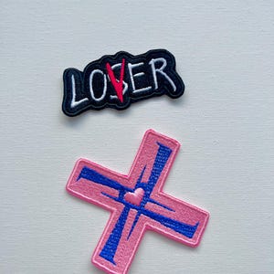 May include: Two embroidered iron-on patches. One is black with white text that reads "LOSER" with a red slash through the "O". The other is pink with a blue cross and a small pink heart in the center.