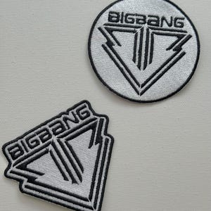 Może przedstawiać: Dwa białe haftowane naszywki z napisem "BIGBANG" i stylizowanym trójkątnym logo w kolorze czarnym.