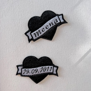 Peut inclure: Deux patchs thermocollants brodés noirs en forme de cœur avec une bannière. Le patch du haut porte le nom "Meena" sur la bannière. Le patch du bas porte la date "29.09.2011" sur la bannière.