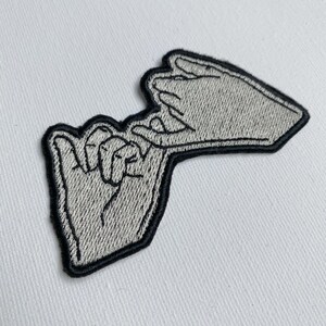 May include: A gray embroidered patch of two hands intertwined, with the fingers of one hand hooked around the fingers of the other hand.