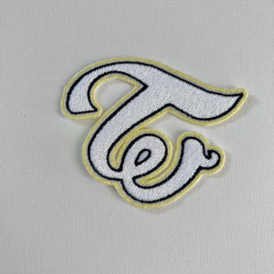 May include: White embroidered patch with the word "Twice" in a stylised script font. The patch has a yellow outline.