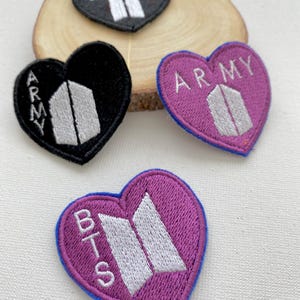 Peut inclure: Trois patchs brodés en forme de cœur avec le texte "BTS" et "ARMY" en lettres blanches sur fond noir et violet. Les patchs présentent le logo BTS, une silhouette blanche stylisée de la lettre "B".