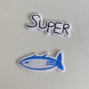 May include: Two embroidered iron-on patches. One patch is white with black outline and says "SUPER". The other patch is white with a blue outline and depicts a fish.