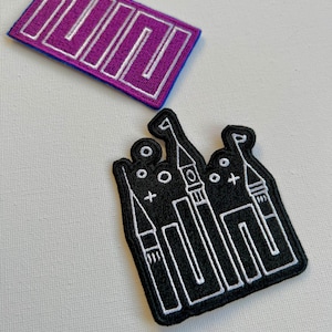 Puede incluir: Dos parches bordados. Uno es morado con contornos blancos de la palabra "IUUD". El otro es negro con contornos blancos de un castillo.