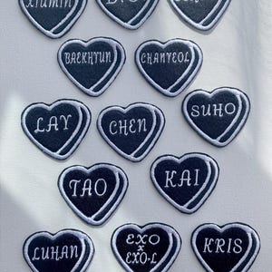 Peut inclure: Douze patchs en forme de cœur noirs avec des contours blancs et du texte blanc. Les patchs présentent les noms des membres du boys band sud-coréen EXO : Xiumin, D.O., Sehun, Baekhyun, Chanyeol, Lay, Chen, Suho, Tao, Kai, Luhan et Kris. Un patch présente le texte « EXO x EXO-L ».