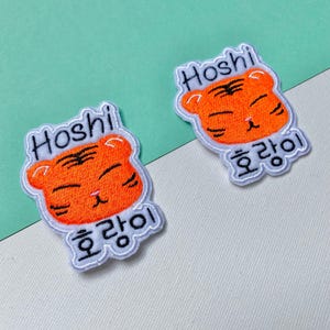 May include: Two iron-on patches featuring a cartoon tiger with the name "Hoshi" and the Korean word "호랑이" (meaning "tiger") written below. The patches are embroidered in orange and white.