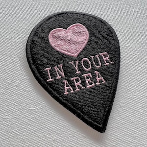 May include: Black felt patch with a pink heart and the text "IN YOUR AREA" in pink thread.