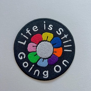 May include: A black and white embroidered patch with a rainbow flower and the text "Life is still going on".