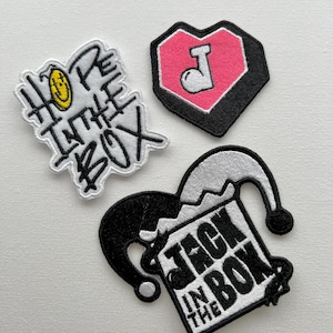 Peut inclure: Trois patchs brodés thermocollants. Un patch est blanc avec du texte noir qui dit "Hope in the Box". Un autre patch est rose avec un contour noir en forme de cœur avec une lettre blanche "J" à l'intérieur. Le troisième patch est noir et blanc avec un chapeau de bouffon et le texte "Jack in the Box".