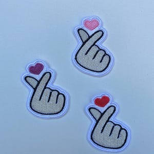 Peut inclure: Trois patchs brodés thermocollants représentant un geste de la main avec un cœur au-dessus. Les patchs sont blancs avec des contours noirs et les cœurs sont roses, rouges et violets.