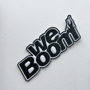May include: Black and white embroidered patch with the text "We Boom" in a bold, stylized font. The "O" in "Boom" is shaped like a flame.