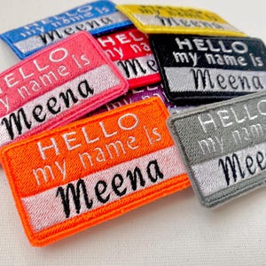 Puede incluir: Un conjunto de parches bordados de colores para planchar con el texto "HELLO my name is Meena".