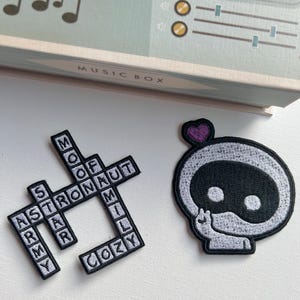 May include: Two embroidered patches. One is a crossword puzzle with the words "astronaut", "cozy", "star", "moon", "family", and "arm". The other is a white and black cartoon astronaut with a purple heart on its head.