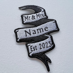 Könnte beinhalten: Schwarzer und weißer gestickter Patch mit einem Banner-Design. Das Banner lautet "Mr. & Mrs. Name Est. 2023".