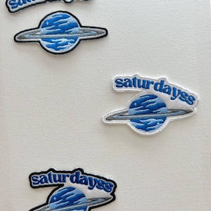 May include: Three embroidered patches with a blue planet and rings, and the text "saturdayss" in a white, blue, and black font.