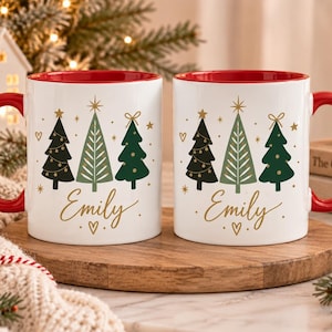 Könnte beinhalten: Zwei weiße Keramiktassen mit roten Henkeln und rotem Innenraum, bedruckt mit drei stilisierten Weihnachtsbäumen in Grün- und Goldtönen, wobei der Name Emily in goldener Schreibschrift unter den Bäumen und kleinen goldenen Sternen steht.