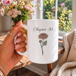 Puede incluir: Una taza de cerámica blanca con el texto negro Chapter 50 sobre una ilustración de líneas de un crisantemo rosa polvoriento con hojas y tallo verdes. La taza tiene un acabado liso y un asa curva en el lado izquierdo para sujetarla.