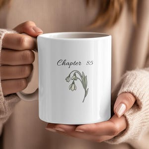 Puede incluir: Taza de cerámica blanca con el texto "Chapter 85" en fuente cursiva, acompañada de una delicada ilustración botánica de flores de campanillas de invierno en verde y blanco. La taza se sostiene con ambas manos.