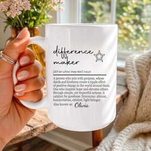 May include: A white ceramic mug featuring black printed text that defines the term difference maker, including a phonetic pronunciation, a descriptive definition, and the name Olivia at the bottom, accompanied by a small star graphic.