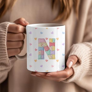 May include: White ceramic mug featuring a patchwork letter "N" design and small heart motifs in multiple colours. The mug is held by a person wearing a beige jumper.