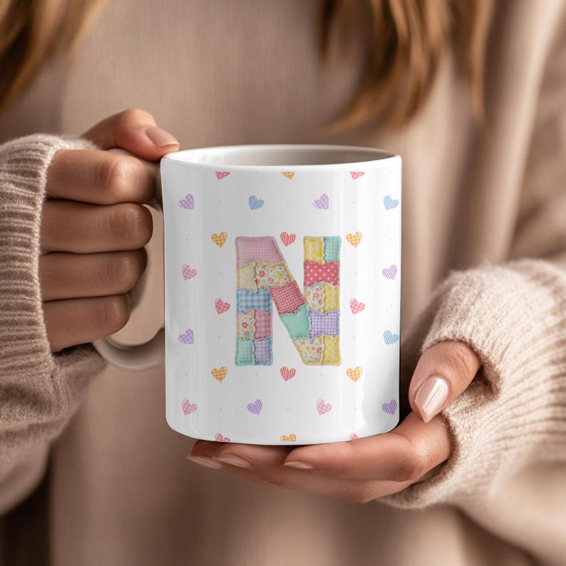 May include: White ceramic mug featuring a patchwork letter "N" design and small heart motifs in multiple colours. The mug is held by a person wearing a beige jumper.