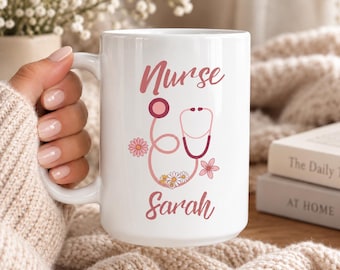 May include: A white ceramic mug featuring the text Nurse and Sarah in a pink script font. The design includes a pink stethoscope graphic intertwined with small daisy flowers in shades of pink, white, and yellow, printed on the front of the mug.