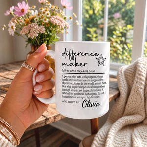 May include: A white ceramic mug featuring black printed text that defines the term difference maker, including a phonetic pronunciation, a descriptive paragraph, and the name Olivia at the bottom, accompanied by a small star graphic.