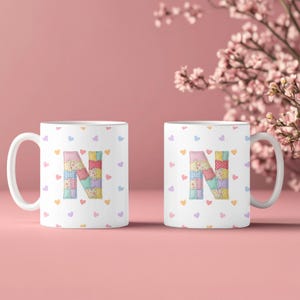 May include: Two white ceramic mugs featuring a patchwork letter "N" in various pastel colors. The mugs are adorned with small heart shapes. The background is a soft pink, with floral accents.