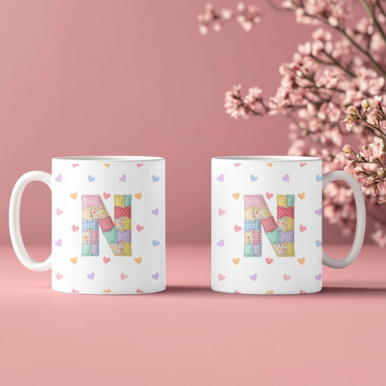 May include: Two white ceramic mugs featuring a patchwork letter "N" in various pastel colors. The mugs are adorned with small heart shapes. The background is a soft pink, with floral accents.