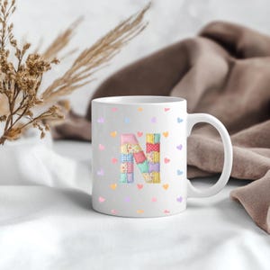 May include: White ceramic mug featuring a patchwork letter "N" in pastel shades. The mug is decorated with small heart patterns in various colours. The mug has a curved handle and is set against a soft, neutral background.
