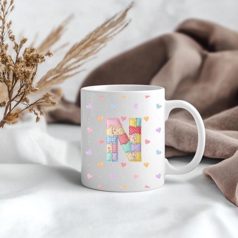 May include: White ceramic mug featuring a patchwork letter "N" in pastel shades. The mug is decorated with small heart patterns in various colours. The mug has a curved handle and is set against a soft, neutral background.