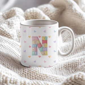 May include: White ceramic mug with a patchwork letter "N" in pastel shades. The mug is decorated with small, colourful hearts. The mug has a large handle and is set against a soft, white, textured background.