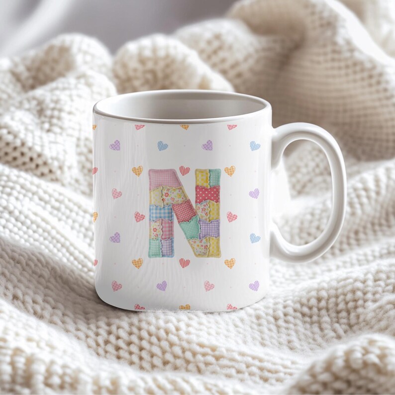 May include: White ceramic mug with a patchwork letter "N" in pastel shades. The mug is decorated with small, colourful hearts. The mug has a large handle and is set against a soft, white, textured background.