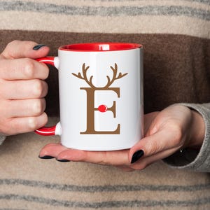 Puede incluir: Taza de cerámica blanca con interior y asa rojos. La taza presenta la letra marrón "E" con cuernos de reno y una nariz roja, perfecta para la temporada navideña.