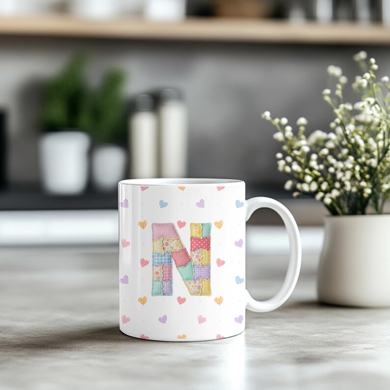 May include: White ceramic mug with the letter "N" in a patchwork design. The letter is made up of different fabric patterns and colours. The mug is decorated with small, colourful heart shapes.