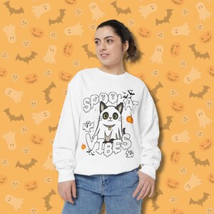 Puede incluir: Sudadera blanca con un diseño de Halloween. El diseño presenta un gato disfrazado de fantasma con las palabras "Spookat Vibes". El fondo es naranja con murciélagos, fantasmas y calabazas.