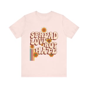Könnte beinhalten: Hellrosa T-Shirt mit dem Schriftzug "SPREAD LOVE NOT HATE" in Retro-Schrift. Das Design beinhaltet Blumen und einen vertikalen Streifen mit grünen, roten und gelben Farben. Ein lässiges Kleidungsstück.