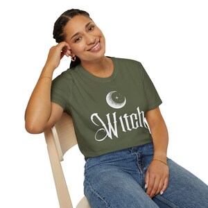 Op de afbeelding: Olijfgroen t-shirt met het woord "Witch" in witte letters. Boven het woord staat een wit maan- en sterontwerp. Het t-shirt heeft een ronde hals.