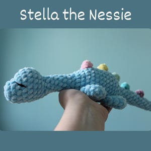 Può includere: Un giocattolo di peluche Nessie all'uncinetto blu con accenti colorati. Il giocattolo è tenuto in una mano.