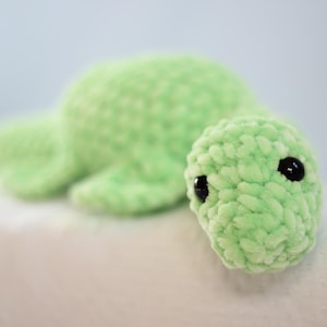 Op de afbeelding: Een groen gehaakt pluche speelgoed in de vorm van een schildpad met zwarte ogen. De schildpad ligt op zijn buik met zijn kop naar de zijkant gedraaid.