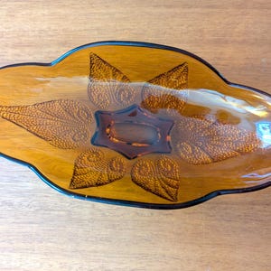 May include: An amber-colored glass serving dish with a leaf design. The dish has a dark brown rim and a central star-shaped indentation. The leaf patterns are embossed, adding texture to the glass.