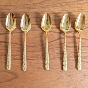 May include: Five gold-colored spoons with an intricate design on the handle. The spoons are arranged in a row on a wooden surface.