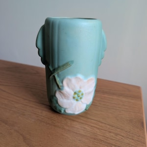 May include: A turquoise ceramic vase with a raised floral design. The vase features a stylized flower with white petals and a green center. The vase has vertical lines and wing-like handles on either side.