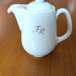 May include: A small, white ceramic teapot with a lid, spout, and handle. The teapot has the initials "FR" in a delicate, green script on the front. The teapot is sitting on a wooden surface.
