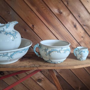 May include: A white ceramic wash set featuring a large pitcher and basin, a chamber pot, a small mug, and a small pitcher. Each piece is decorated with a light blue ornate scrollwork pattern and scalloped edges, creating a cohesive set.