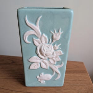 May include: A rectangular, light blue ceramic vase with a white, raised floral design. The design features a rose and leaves. The vase is displayed on a wooden surface.